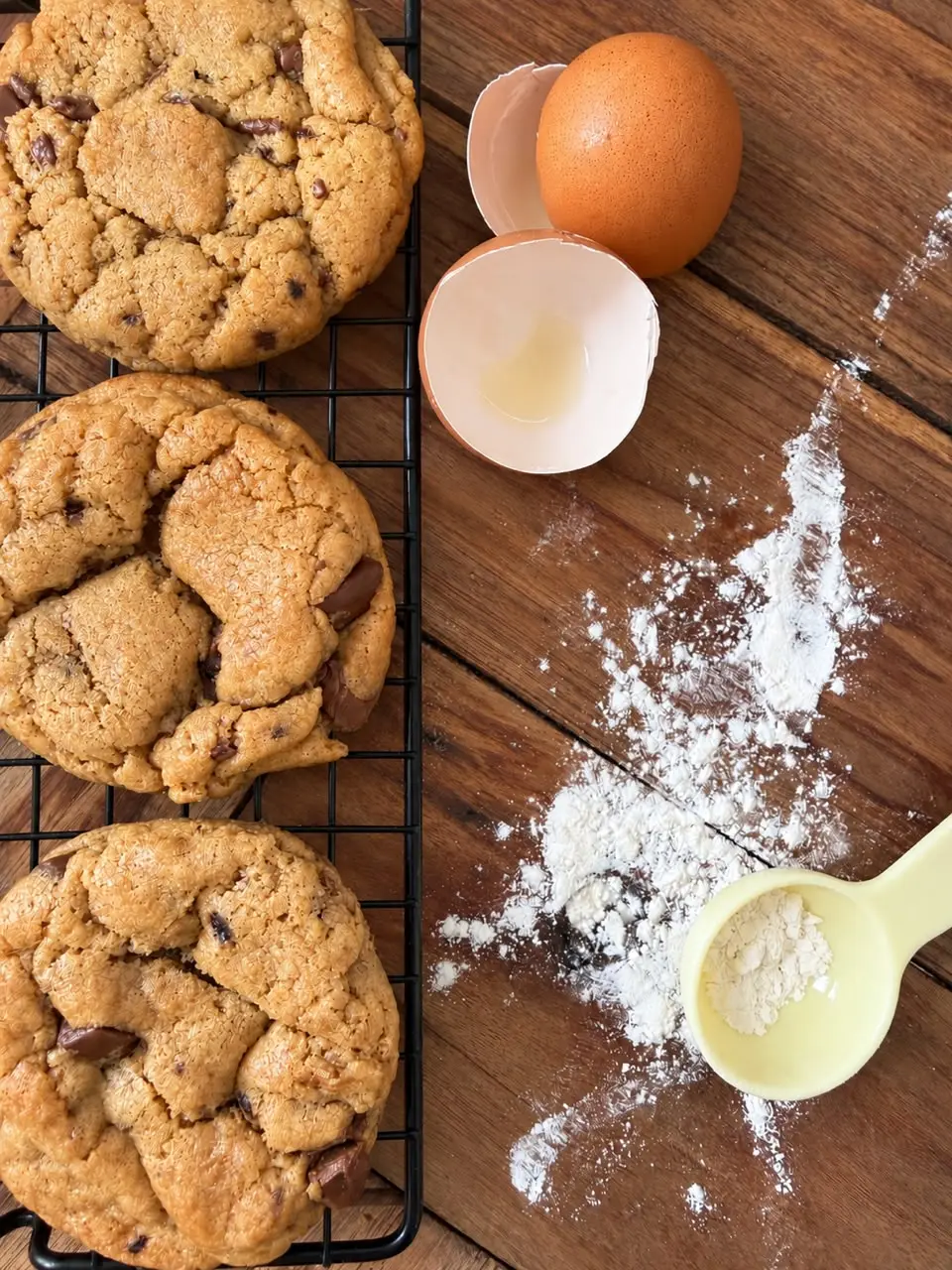Three thick choc chip cookies cooling on a wire rack beside a cracked brown egg and a scattering of flour on a wooden table